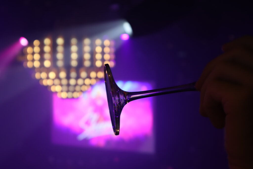 A close-up view of an inverted champagne glass held against vibrant pink and purple stage lighting aboard a Virgin Voyages ship.