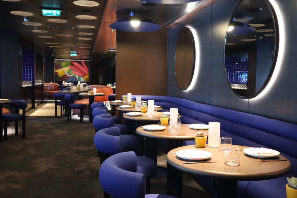 The interior of Pink Agave, Virgin Voyages’ modern Mexican restaurant, featuring blue seating, round tables and ambient low lighting.