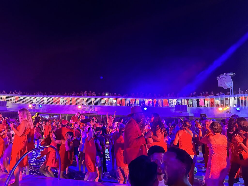 A vibrant Scarlet Night pool party onboard Virgin Voyages, with Sailors dressed in red dancing under colourful lighting on the deck.