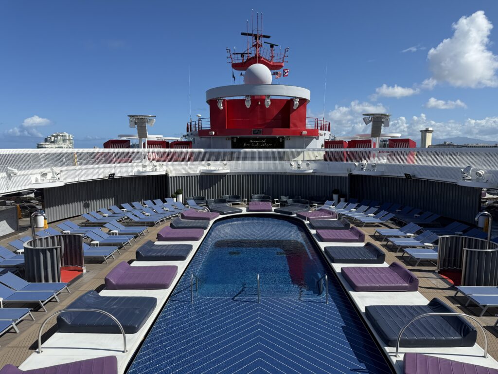 The main pool deck onboard Virgin Voyages’ Valiant Lady, featuring sun loungers, purple cushions, and a central pool under clear blue skies.