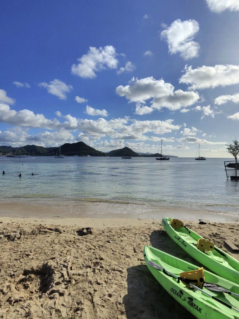 Green kayaks on a sandy beach in St Lucia with calm Caribbean water and sailboats during a Virgin Voyages Shore Thing excursion