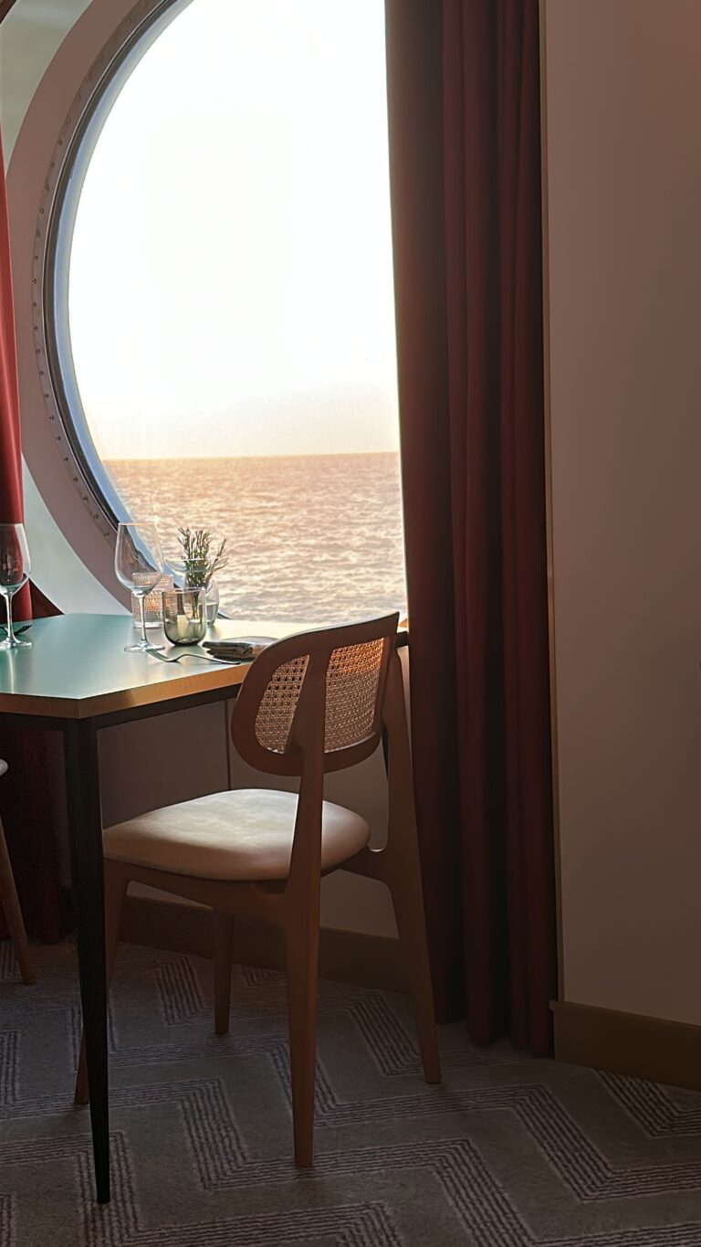 Interior of Virgin Voyages Extra Virgin restaurant featuring an ocean-view table beside a large circular window overlooking the sea during a cruise voyage.