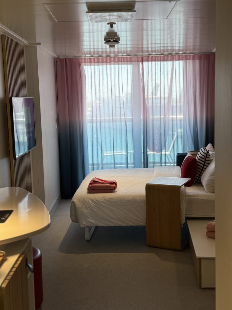 Interior of a Virgin Voyages Sea Terrace cabin featuring a modern bed, colourful décor, floor-to-ceiling balcony doors and ocean views.