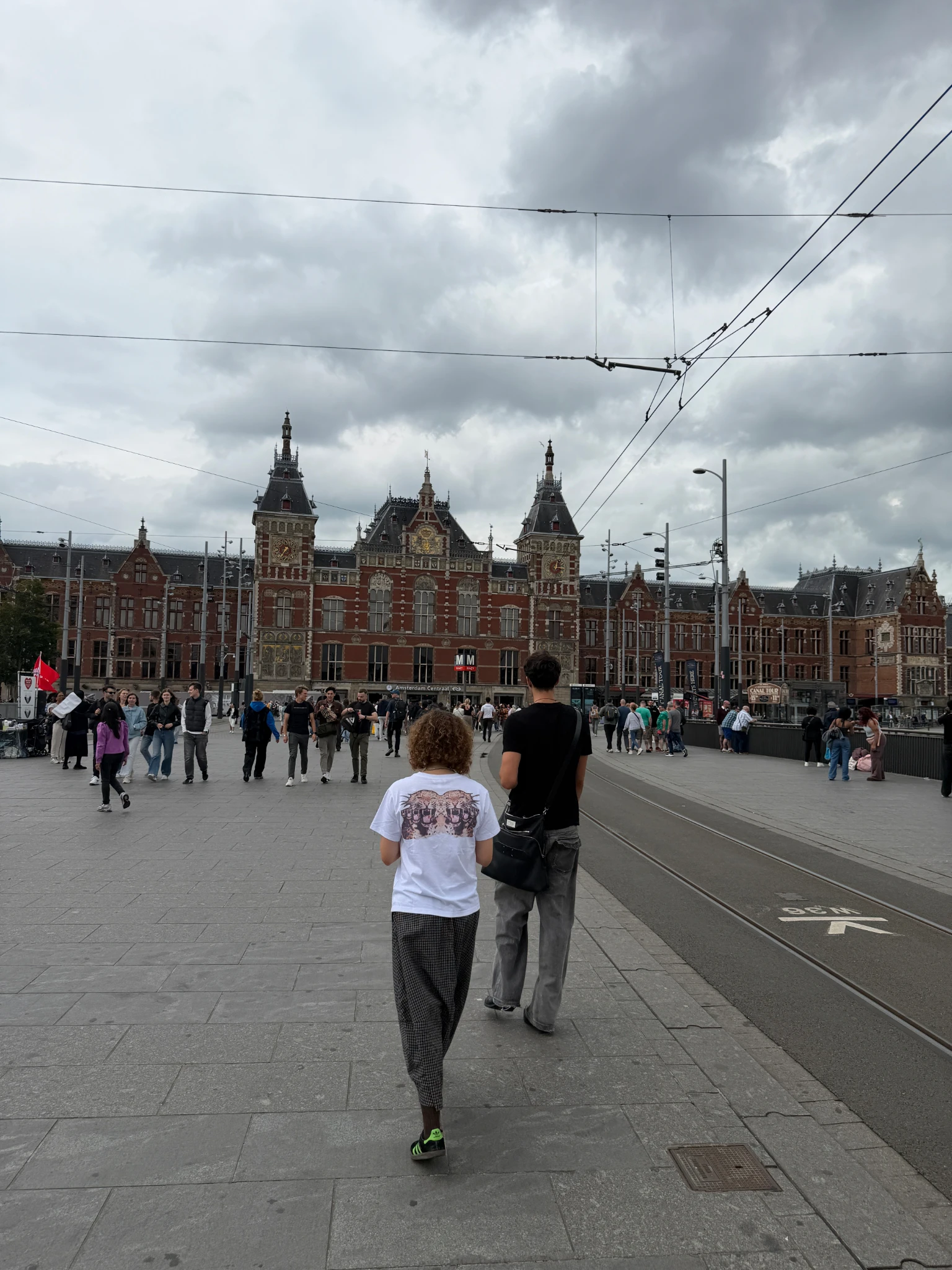 Amsterdam Centraal Station viewed from the waterfront plaza with pedestrians, tram tracks and historic Dutch architecture during a Virgin Voyages visit to Amsterdam.