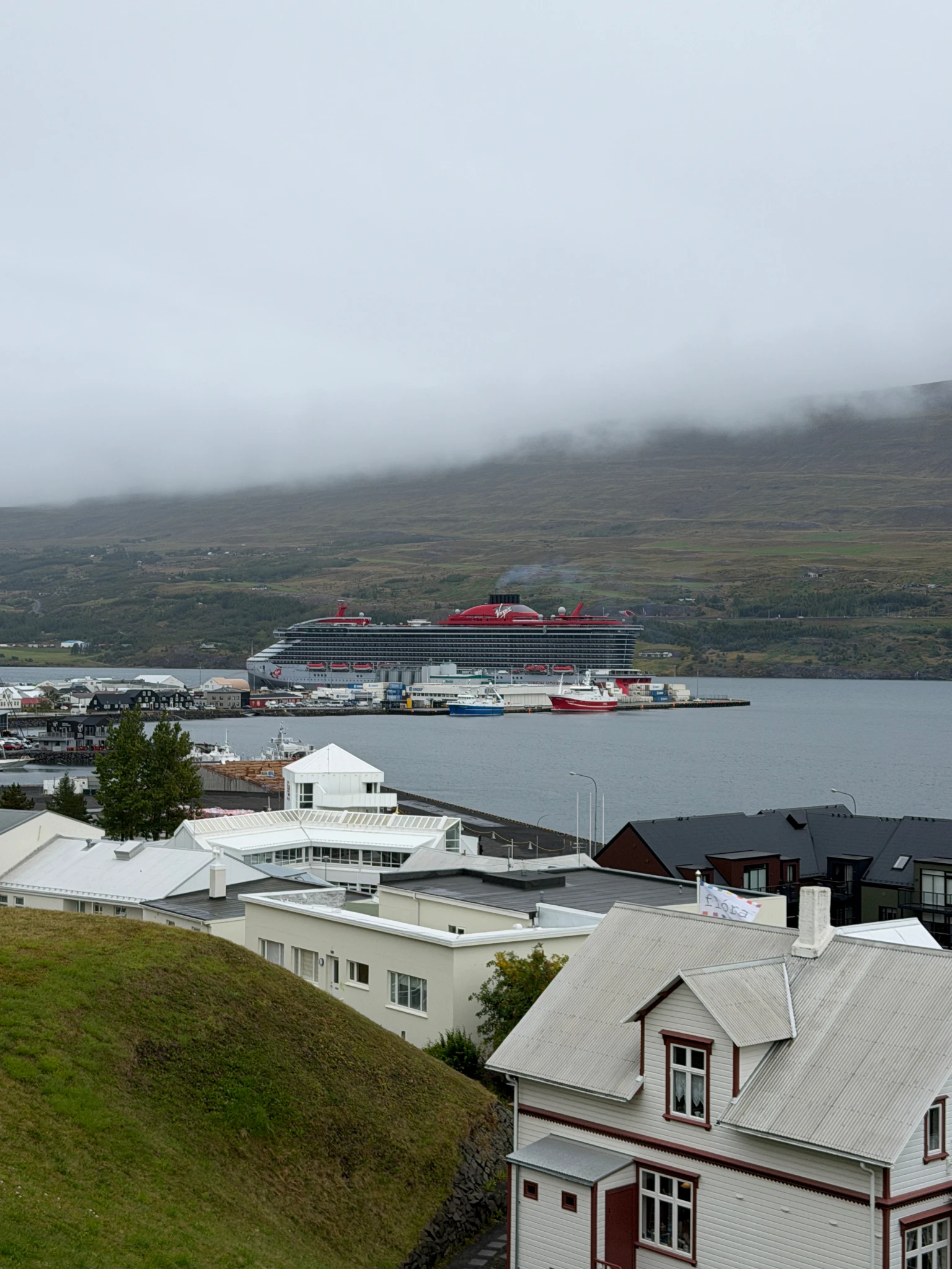 Virgin Voyages cruise ship docked in Akureyri Harbour with Icelandic mountains, low clouds and waterfront buildings overlooking Eyjafjörður fjord.