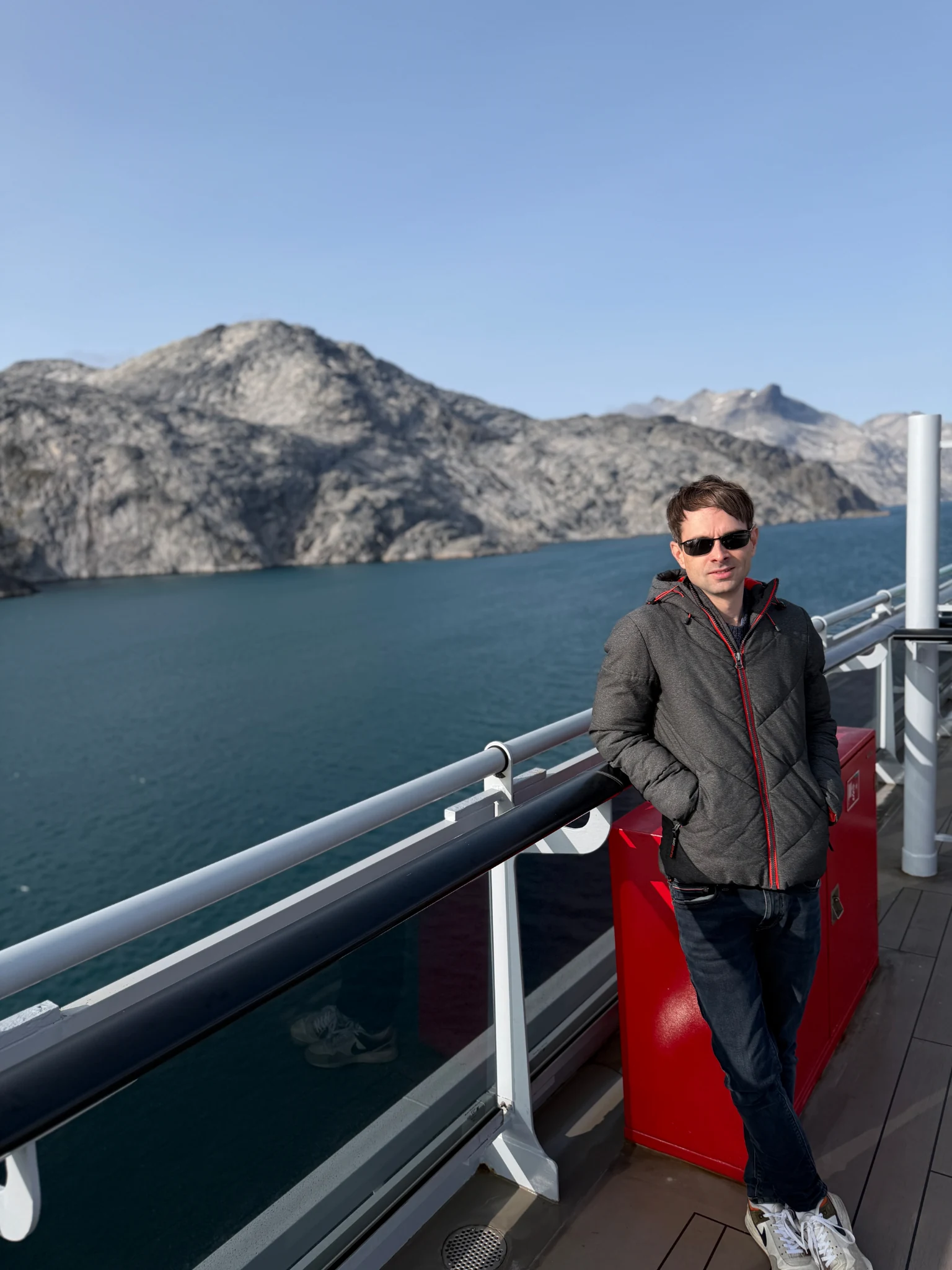 Daniel Griese beside a Greenland flag with fjord and mountain scenery during a Virgin Voyages sailing