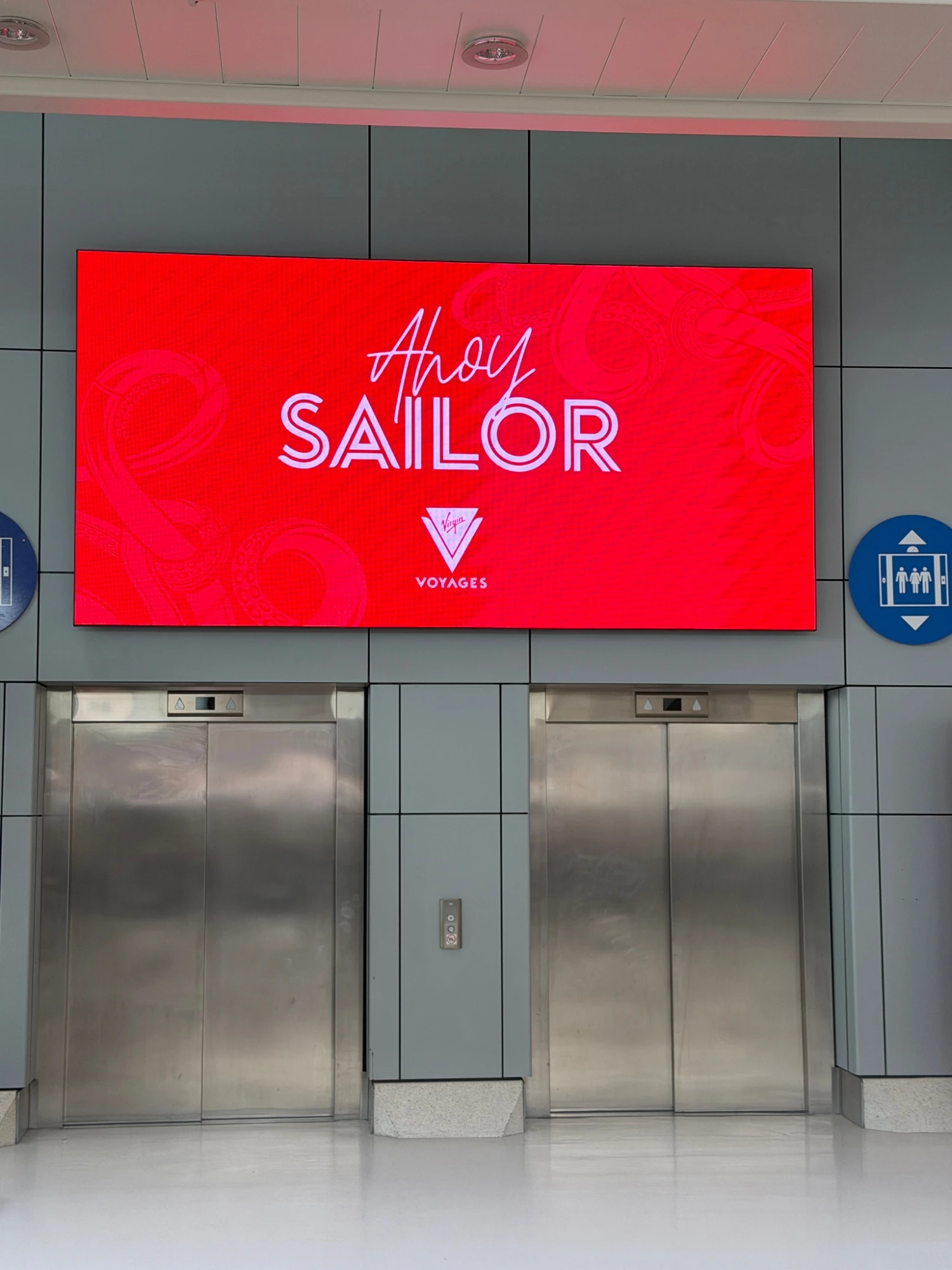 Virgin Voyages Ahoy Sailor digital welcome screen displayed above lifts inside Portsmouth Cruise Terminal before embarkation.
