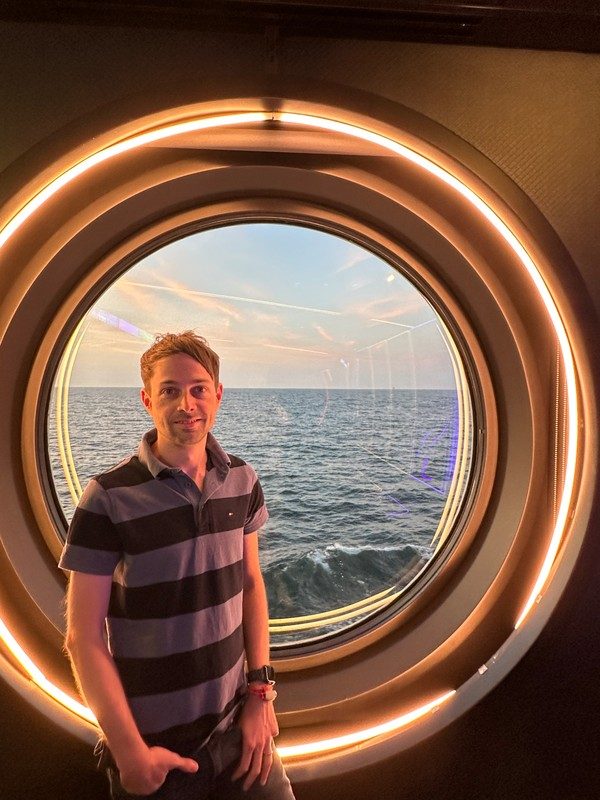Daniel Griese onboard a Virgin Voyages cruise ship, standing in the casino beside a circular porthole window overlooking the open ocean at sunset