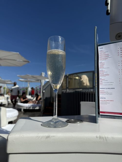 Glass of champagne on an outdoor sun deck table aboard a Virgin Voyages ship, with a drinks menu, loungers, parasols and a clear blue sky in the background.